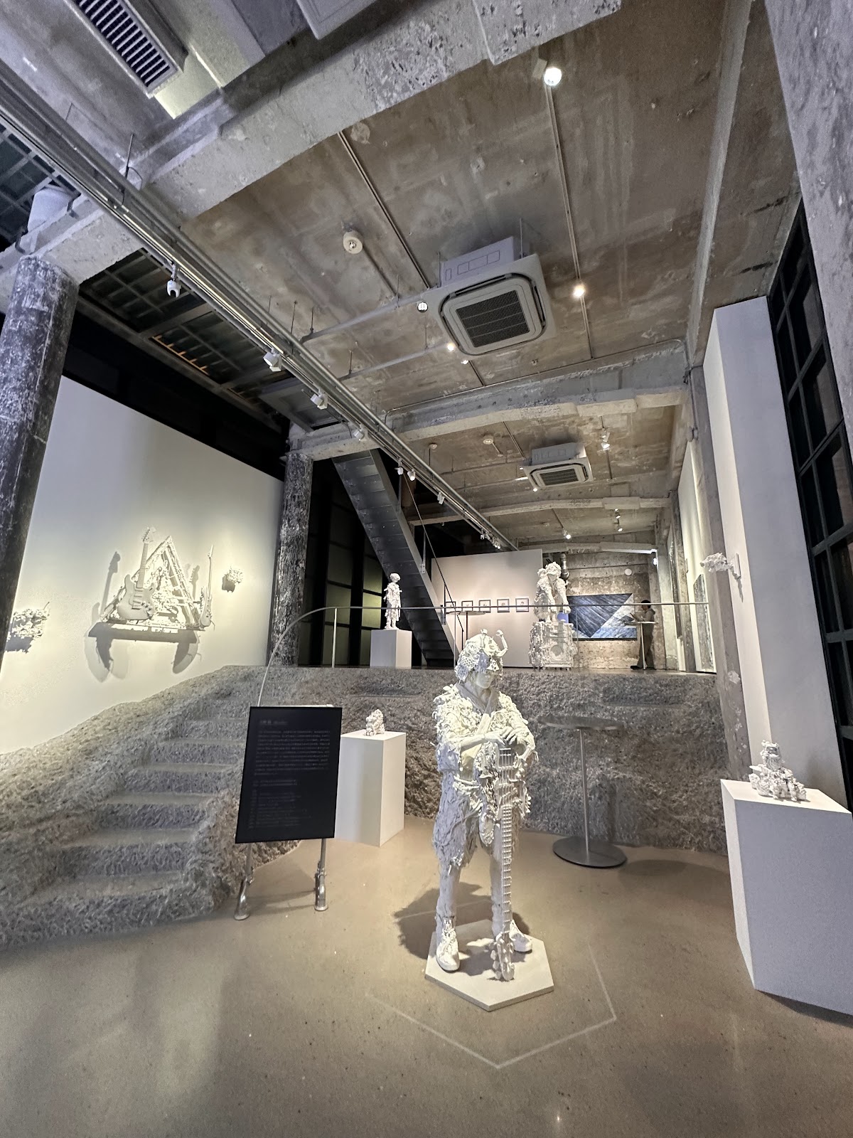 コンクリートの天井と白い壁の展示室。中央に白い人型の彫刻が立ち、壁にはギターを組み込んだ白い立体作品。左手に岩のような階段。